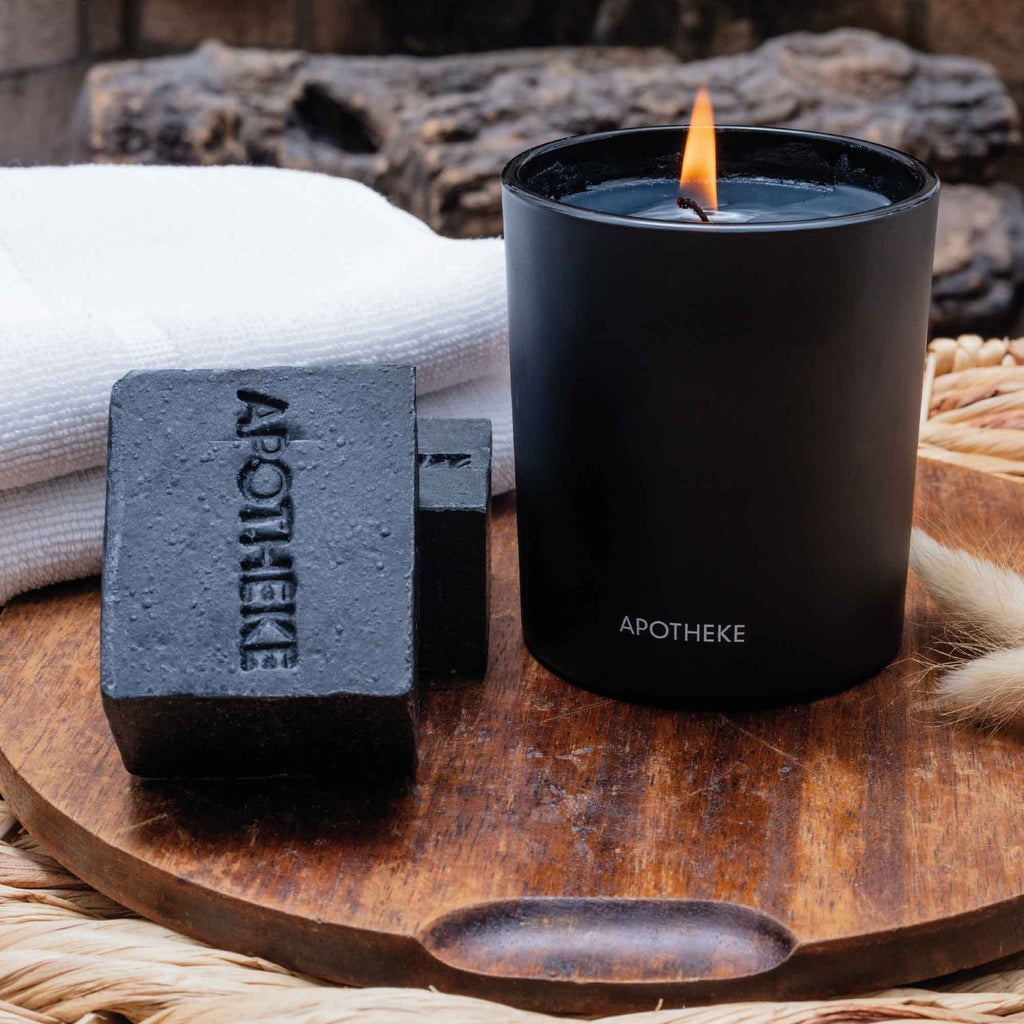 A lit black candle sits in a holder labeled Apotheke on a wooden tray beside two Apotheke Charcoal Bar Soaps. The woody fragrance fills the air, enhanced by a folded white towel and a wicker basket that create a cozy backdrop.