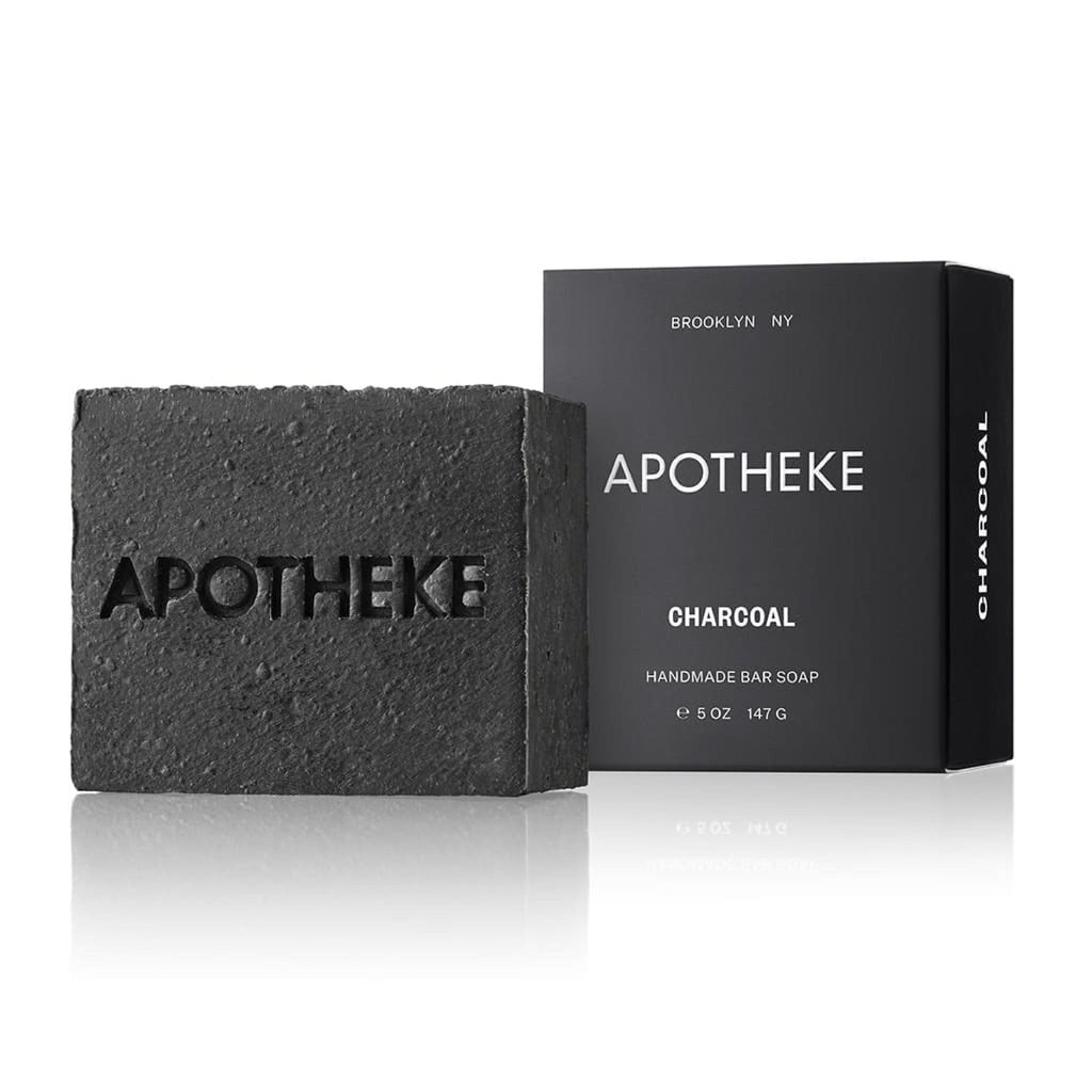 The Apotheke Charcoal Bar Soap, rectangular and black, is embossed with APOTHEKE and emits a subtle woody aroma. It rests next to its chic black box labeled APOTHEKE and CHARCOAL, emphasizing its plant-based ingredients and product details.