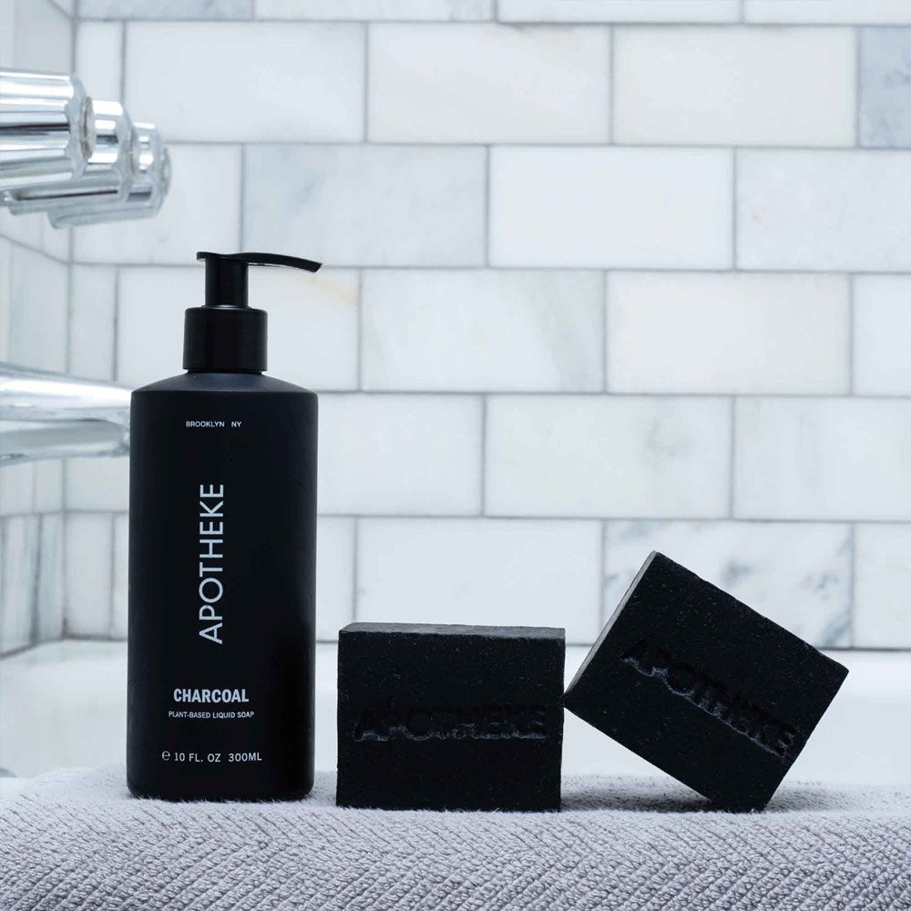 Apothekes Charcoal Liquid Soap bottle, infused with natural botanicals, is displayed with two black soap bars on a textured surface in a white-tiled bathroom. A shiny faucet appears partially on the left.