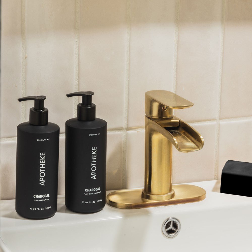 Two bottles of Apotheke - Charcoal Lotion with pump tops elegantly rest on a white sink beside a modern gold faucet. The beige tile background enhances the neutral tone of this plant-based setting.