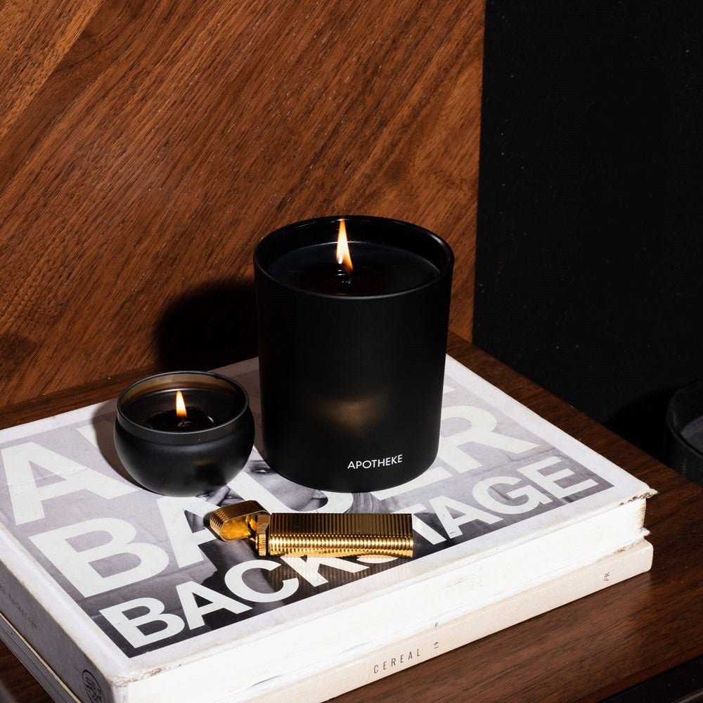 An Apotheke Charcoal Mini Tin 2oz Candle is lit atop a stack of books with visible titles. A gold lighter rests nearby on the wooden surface, contrasting against the dark charcoal-hued wall in the background.