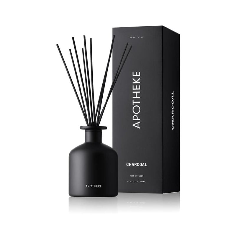 The Apotheke - Charcoal Reed Diffuser features a black bottle with reeds standing next to a matching box. Labeled Apotheke and Charcoal, it offers a modern aroma of cedarwood and sandalwood through its rich essential fragrance oils.