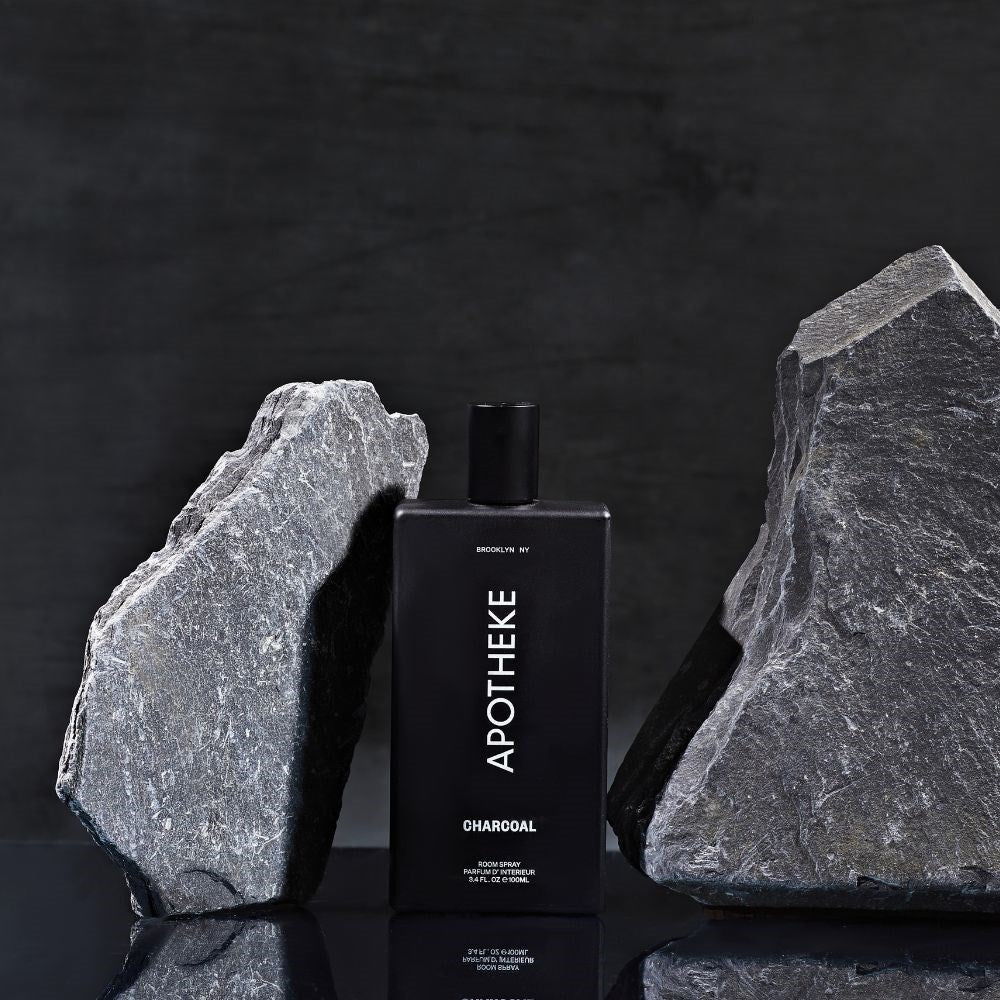 A bottle of Apotheke Charcoal Room Spray, similar to luxury liquid soap, stands between large gray rocks against a dark backdrop. The rectangular bottle has a black cap and white lettering, subtly hinting at its woody fragrance through its elegant design.