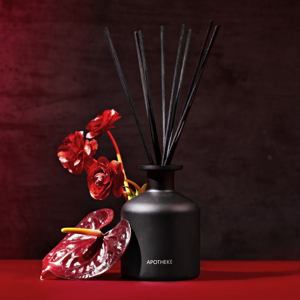 An Apotheke - Charcoal Rouge Reed Diffuser with black reeds sits on a red surface next to red anthurium flowers. The dark background accentuates the allure of the Apotheke diffuser and its captivating fragrance oils.