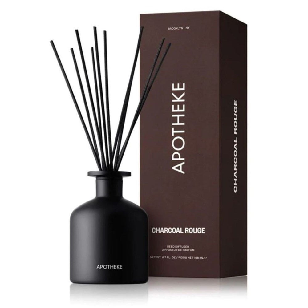 A black Apotheke reed diffuser bottle labeled APOTHEKE is placed beside its matching dark brown CHARCOAL ROUGE box, adorned with BROOKLYN, NY. It features several long black reeds infused with premium fragrance oils.