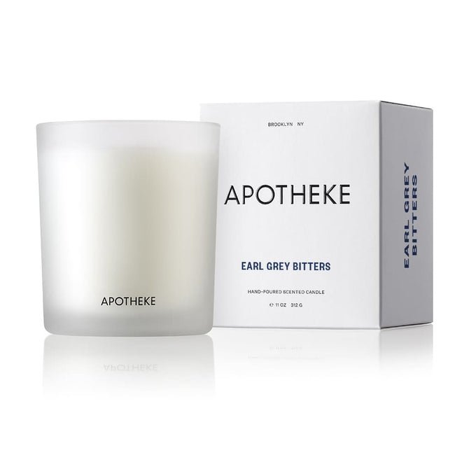 A frosted glass container displays the Apotheke - Earl Grey Bitters 10.5oz Candle, made from a soy wax blend. Next to its box with a minimalistic black-on-white design, it hints at citrusy bergamot.