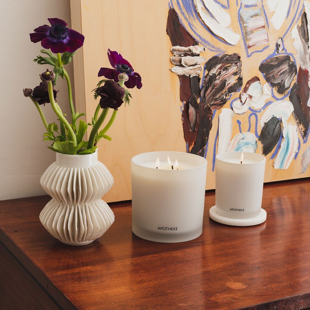 A white ribbed vase with purple flowers sits beside an Apotheke - Earl Grey Bitters 3-Wick Candle on a wooden surface. Its soy wax blend brings serenity, while an abstract painting in the background adds a vibrant burst of color.