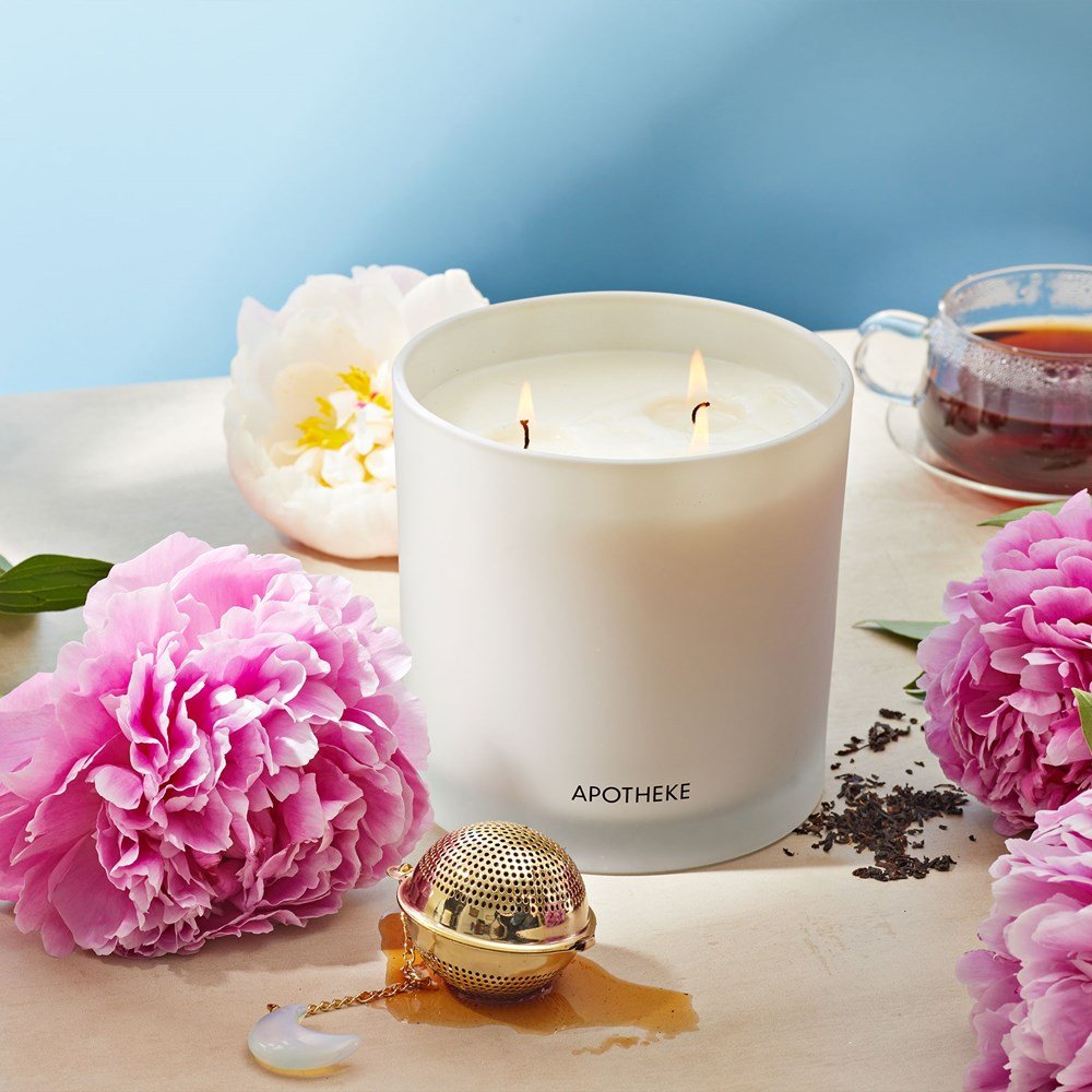 An Apotheke Earl Grey Bitters 3-Wick Candle with a soothing soy blend sits among pink and white peonies. Nearby is a gold tea infuser adorned with a moon charm and an Earl Grey Bitters tea cup, set against a light blue backdrop for a serene ambiance.