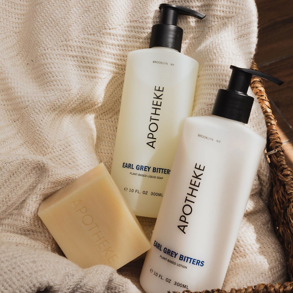 Two Apotheke - Earl Grey Bitters products, a liquid soap and lotion, are elegantly placed on cream-colored knitted fabric. A matching bar soap completes the scene as a woven basket peeks out in the background.