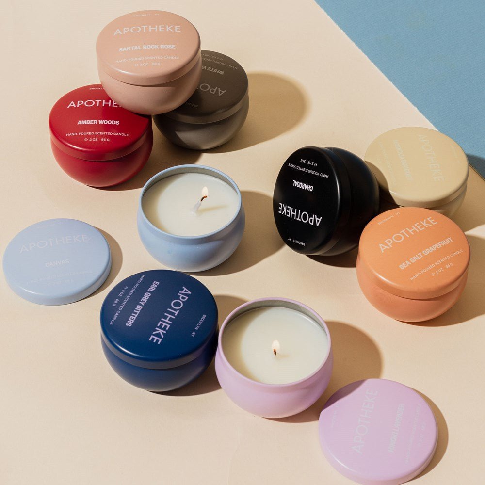 Apotheke offers a collection of round soy wax blend candles in pink, blue, red, and beige with visible wicks. Each tin lid is labeled with scents like Amber Woods, Earl Grey Bitters, and Sea Salt Grapefruit on a neutral background.