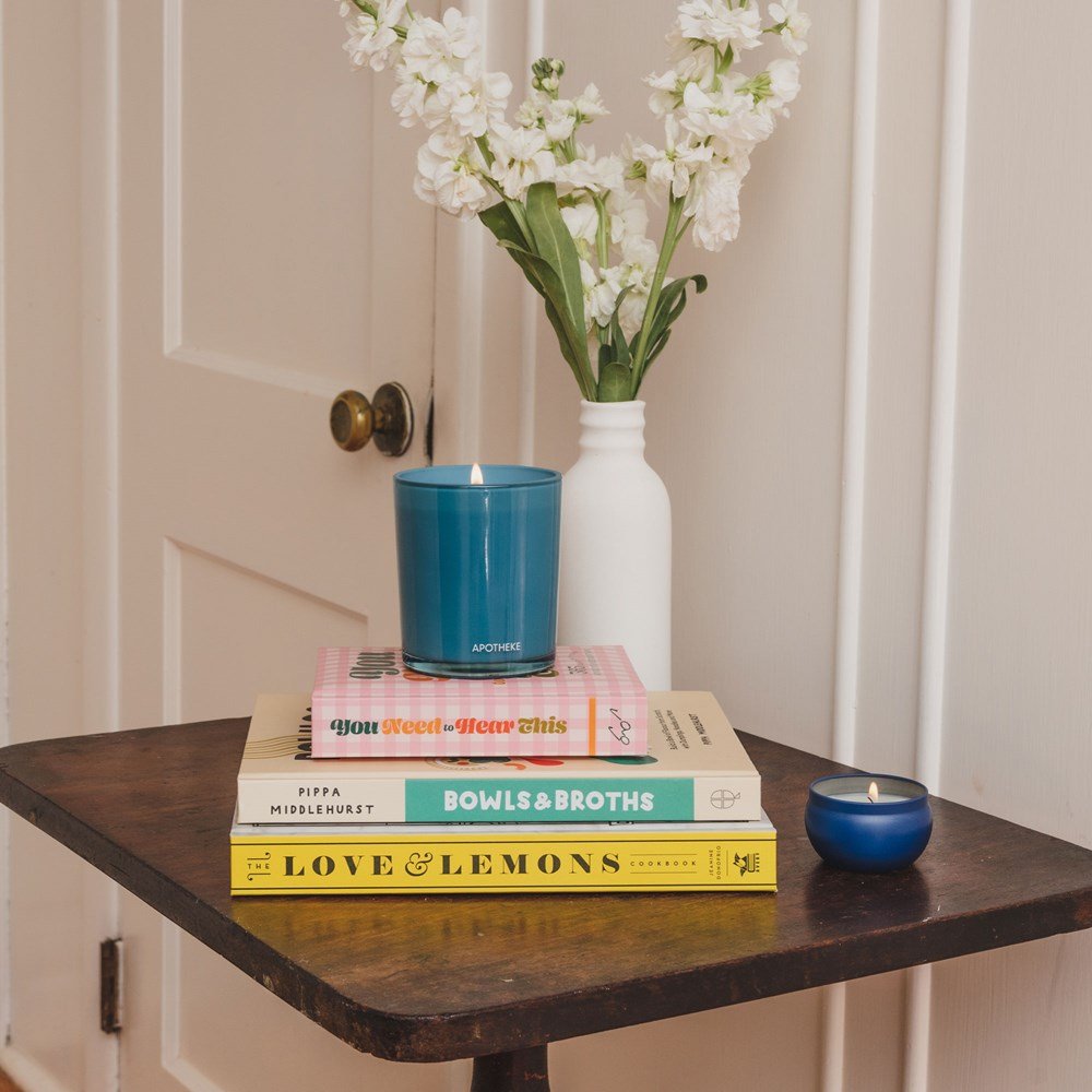 A wooden table holds a stack of three cookbooks and two candles—an Apotheke Earl Grey Bitters Mini Tin 2oz Candle in blue soy wax blend atop the books and a smaller candle beside it. A white vase with white flowers is at the back, all against a light-colored wall.