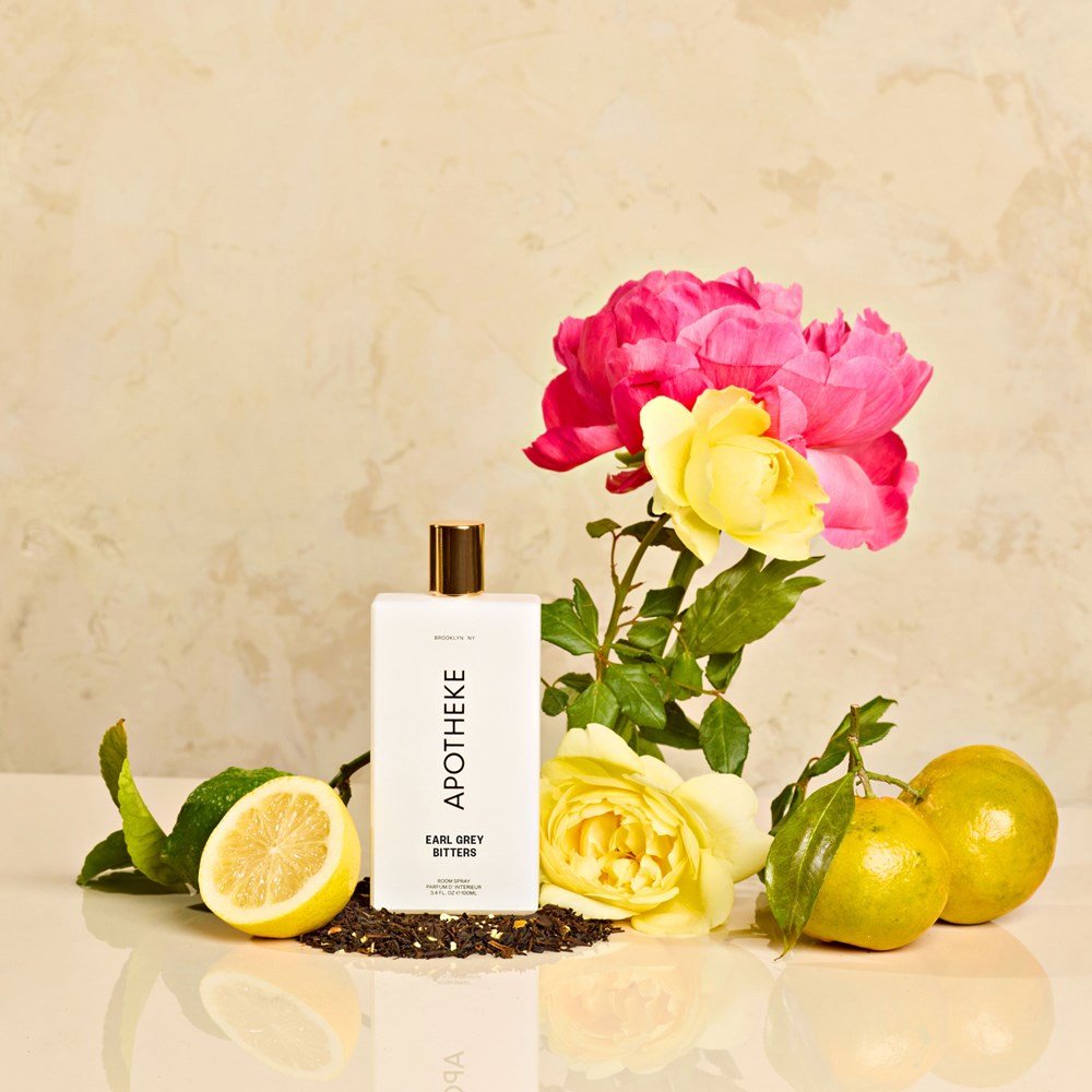 A bottle of Apotheke - Earl Grey Room Spray is set among a cut lemon, black tea leaves, two yellow roses, a pink flower, and three whole lemons on a light marble background, capturing the essence of fine fragrance oils.