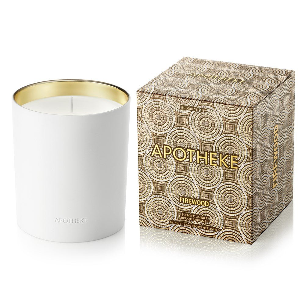 A metallic gold-rimmed jar holding a white Apotheke Firewood 12.5oz Candle rests beside a geometric patterned box featuring APOTHEKE and FIREWOOD in gold lettering, enhancing the woody fragrance ambiance.