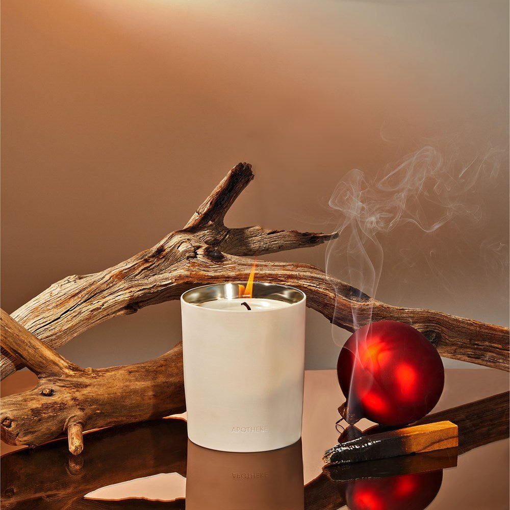 The Apotheke - Firewood 12.5oz Candle in a white ceramic holder is lit on a reflective surface beside a red ornament, driftwood, and an incense stick emitting a woody scent. The backdrop features warm beige and brown gradients.