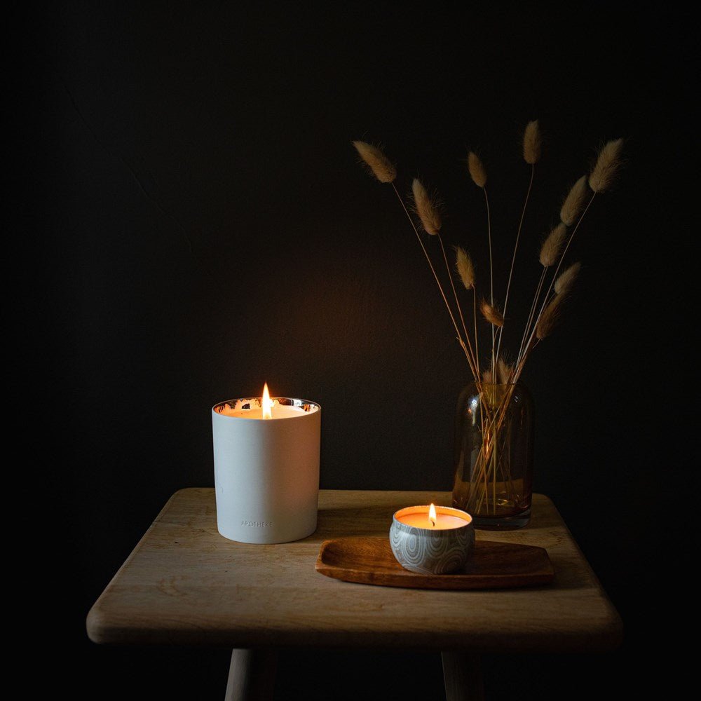 A serene scene features two lit candles on a wooden stool: the Apotheke - Firewood 12.5oz Candle in a tall white holder with a woody fragrance, and another candle nestled in a patterned bowl on a wooden tray. A brown vase displays dried grass against the dark backdrop.