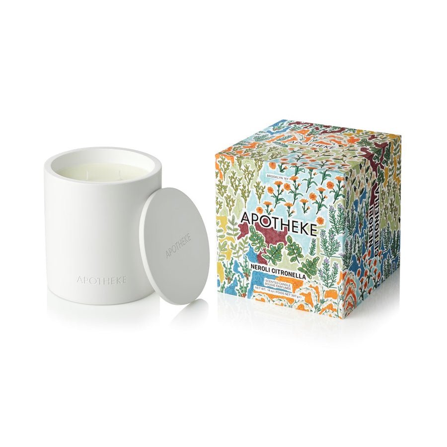 A white luxury Apotheke 2 Wick Candle in “Neroli Citronella” rests beside its vibrant floral box. Both feature bold branding and botanical art. Crafted from a soy wax blend, the candle comes with a matching lid for a premium look.