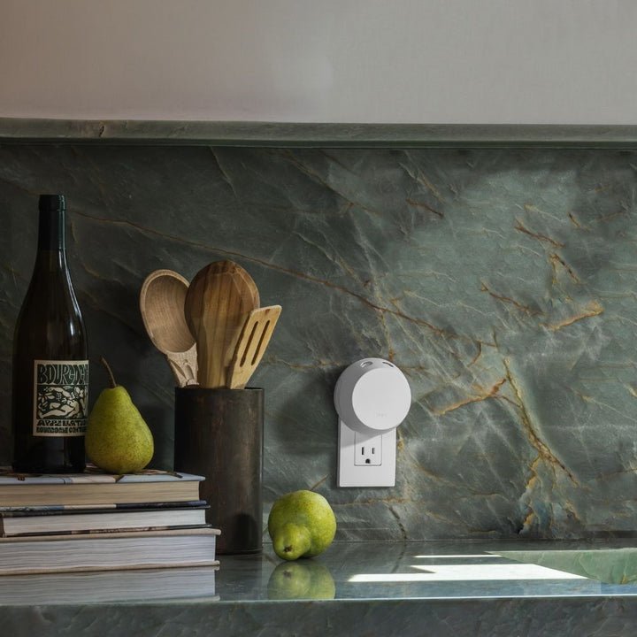 A countertop with green marble showcases an Apotheke - Pura Smart Fragrance Diffuser Set beside a bottle, stacked books, a black container with wooden utensils, two pears, and a plugged-in smart home device. Easily customize your atmosphere using smartphone control for the perfect ambiance.