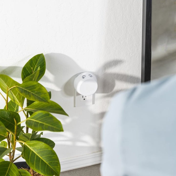 A round, white Apotheke - Pura Smart Fragrance Diffuser Set is plugged into the wall, casting a shadow on the white surface. Nearby, a green plant adds natures touch. A shoulder is visible in the foreground, highlighting fragrance customization through smartphone control by Apotheke.