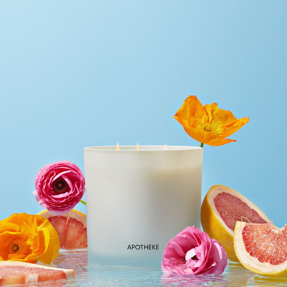 A lit Apotheke - Sea Salt Grapefruit 3-Wick Candle, surrounded by vibrant orange and pink flowers with sliced grapefruit, embodies its citrus aroma. The light blue background enhances this fresh and serene fragrance.