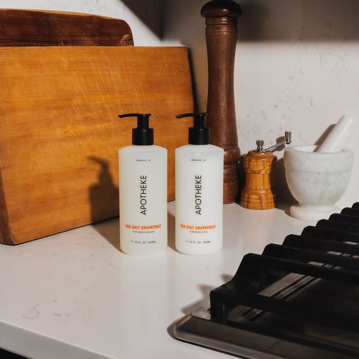 Two bottles of Apotheke Sea Salt Grapefruit lotion are on a kitchen countertop next to a large wooden cutting board, a pepper grinder, a small wooden salt shaker, and a white mortar and pestle, near the stove.