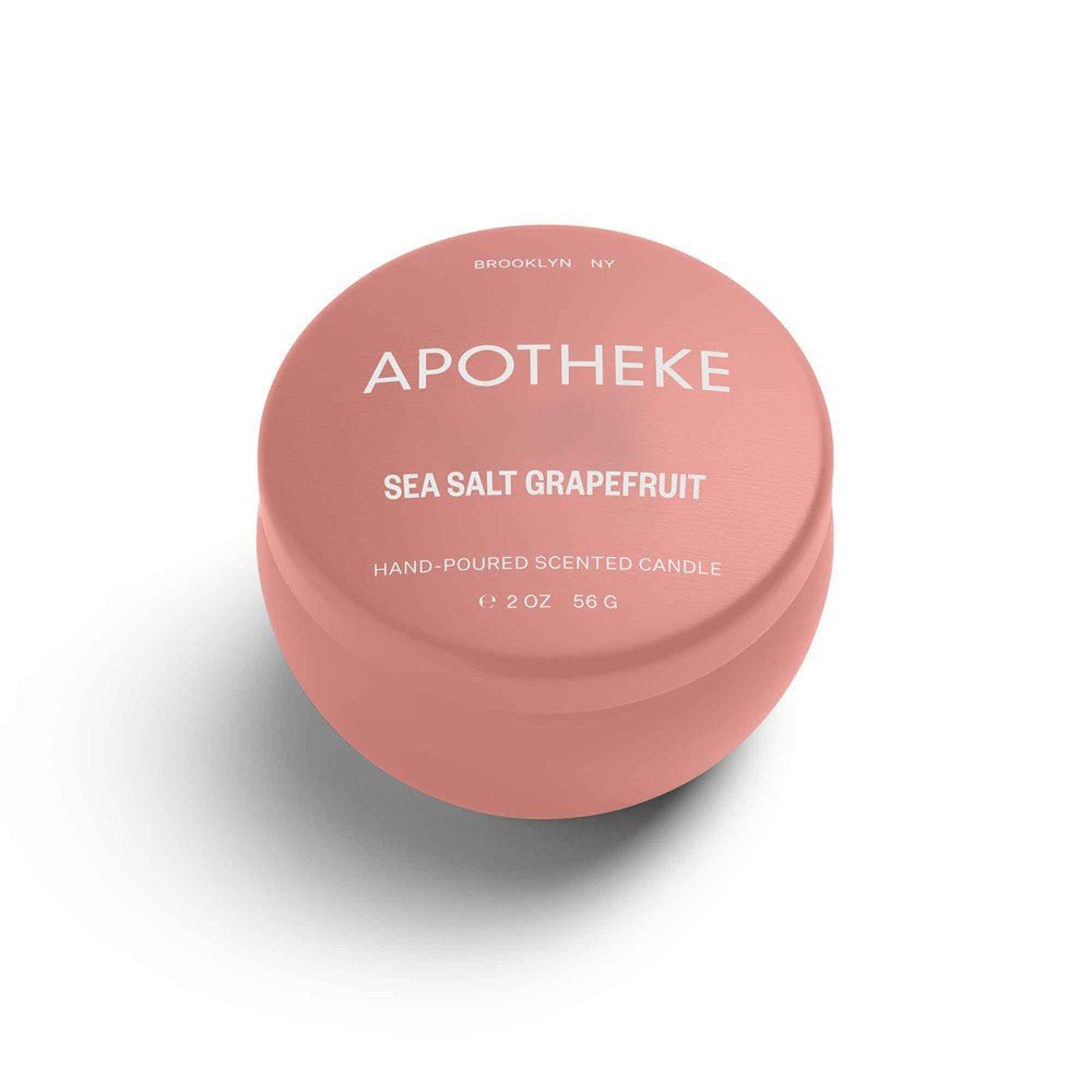 A round, coral tin features the text APOTHEKE - Sea Salt Grapefruit Mini Tin 2oz Candle and emits a refreshing citrus scent. It notes Hand-Poured Soy Wax Candle, weighs 56 g, and mentions Brooklyn NY. The background is crisp white.