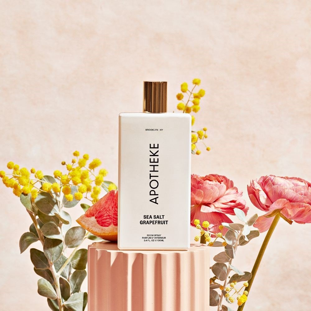 An Apotheke - Sea Salt Grapefruit Room Spray rests on a pink pedestal, releasing its citrus aroma. Its encircled by pink flowers, yellow blooms, and a grapefruit slice set against a soft textured backdrop.