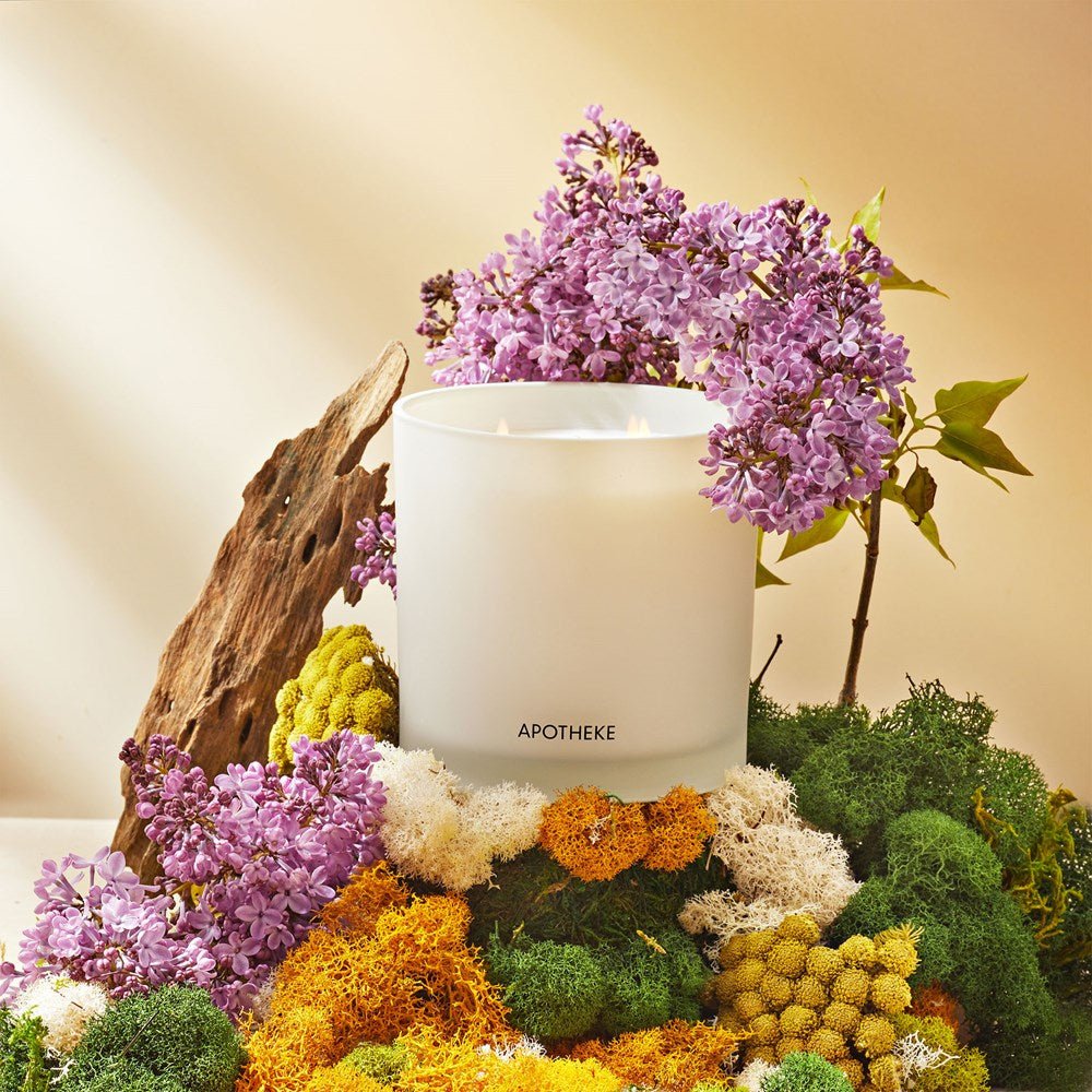 A white Apotheke - White Vetiver 3-Wick Candle crafted from premium soy wax is surrounded by vibrant moss, yellow and white flowers, and purple blooms, all artfully arranged on a wooden surface against a softly lit background.