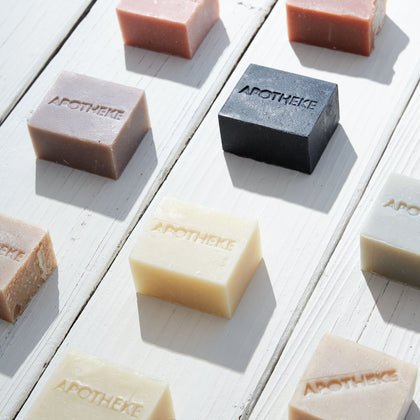 Apothekes White Vetiver Bar Soap, a rectangular, plant-based soap with the brand name engraved, features essential oils. Available in pink, black, beige, and cream hues, it is elegantly placed on a sunlit white wooden surface casting gentle shadows.