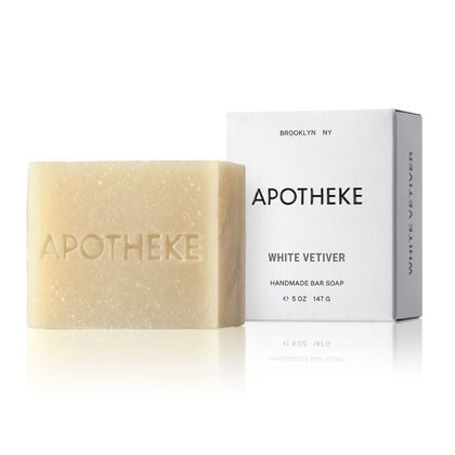 A rectangular APOTHEKE engraved bar soap sits next to its packaging. The white box with black text states Apotheke - White Vetiver, identifying it as a 5 oz (147 g), plant-based, handmade soap from the brand Apotheke.