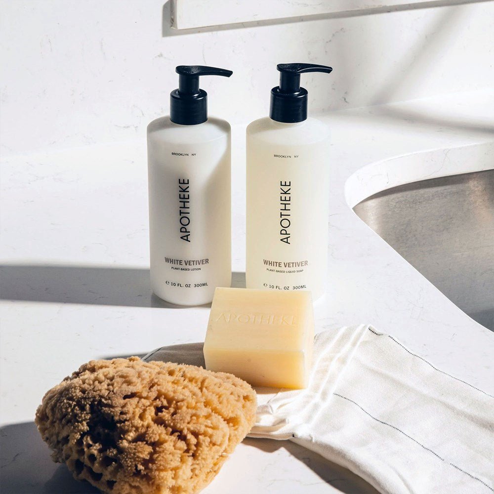 Two Apotheke White Vetiver lotion bottles with pump tops sit on a white countertop. A bar of soap labeled Apotheke rests on a cloth, accompanied by a natural sea sponge near the sink. Infused with plant-based ingredients, these create gentle elegance as natural lighting casts soft shadows.