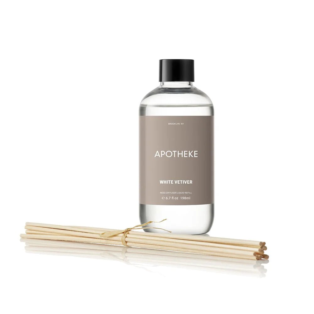 A clear bottle labeled Apotheke - White Vetiver Reed Diffuser Refill sits next to reed sticks on a white background. The bottle, with a black cap and light brown label featuring white text, promises the calming allure of its fragrance oils.