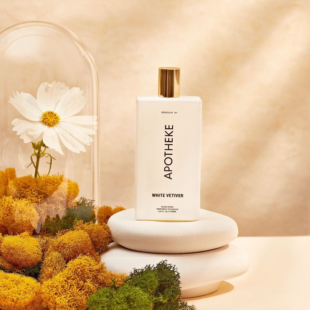 A bottle of Apothekes White Vetiver Room Spray is placed on white circular stones amidst yellow and green moss. A white flower in a glass dome rests on the left, set against a warm, neutral backdrop.