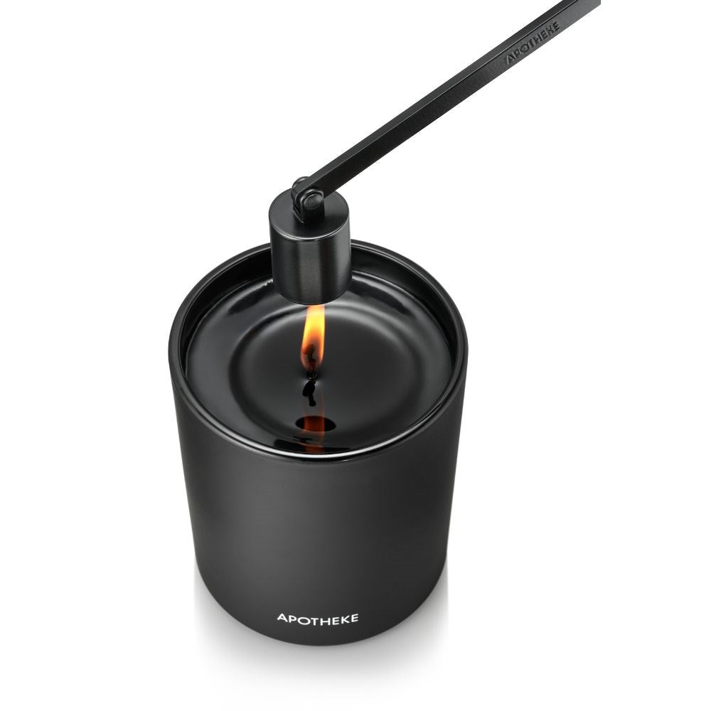 A black candle labeled Apotheke flickers with a lit flame amid melted wax. The Apotheke - Wick Snuffer Black is nearby, ready to extinguish the flame. Its sleek, matte finish adds elegance to any setting.