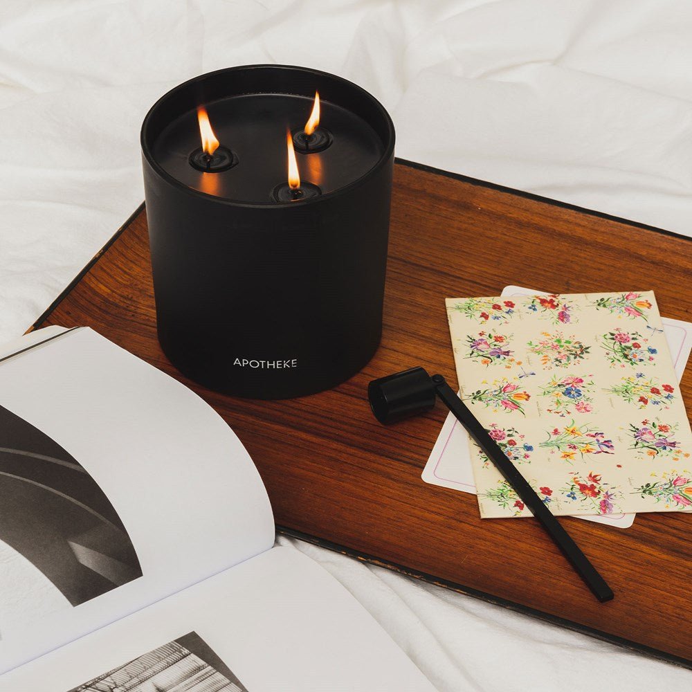 A black three-wick candle labeled APOTHEKE burns on a wooden tray, paired with a floral-patterned card and an Apotheke Wick Snuffer in Black. Nearby, an open book featuring monochrome images lies gracefully on the white bedding.