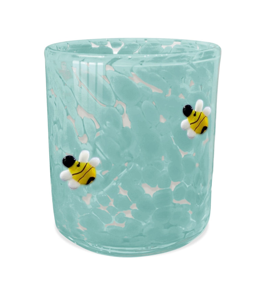 The Aqua De Soi - Bees Knees 12 oz Confetti Candle is a light blue, marbled-textured glass adorned with two decorative bees featuring yellow bodies, black stripes, and white wings. Resembling a whimsical honeycomb, it has a subtle gloss for a shiny finish.