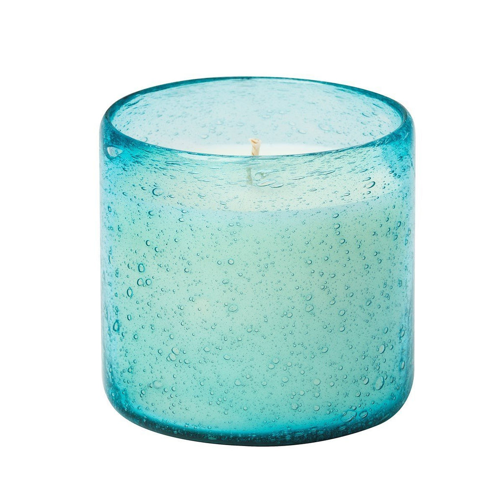 The Aqua De Soi - Blue Agave 8oz Effervescent Candle rests in a blue glass holder with a bubble-like texture, its cylindrical shape cradling the artisan candles unlit wick, ready to release hints of Blue Agave Blossom.