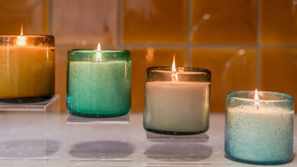 Four lit Aqua De Soi Melon Mojito candles in multicolored glass holders rest on raised platforms. Their warm glow contrasts with a blurred, orange-tiled background, creating a cozy atmosphere that invites you to imagine their soothing fragrance notes.