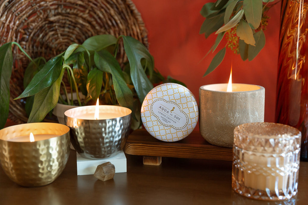 The Aqua De Soi Sage and Vetiver 14oz Round Hammered Bowl Candle is artistically arranged on a wooden surface with sage leaves and a wicker wall decoration. A small crystal nearby enhances the cozy ambiance.