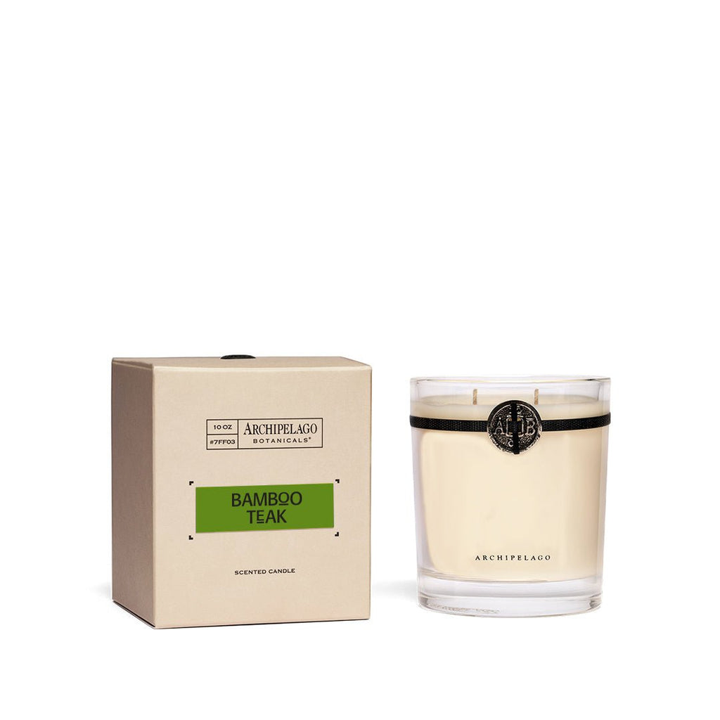 The Archipelago - Bamboo Teak Credenza 10oz Candle, set in a glass holder, exudes a woodsy aroma. It sits beside a beige box made of sustainable materials, featuring a green label. Both the box and candle display the Archipelago branding prominently.