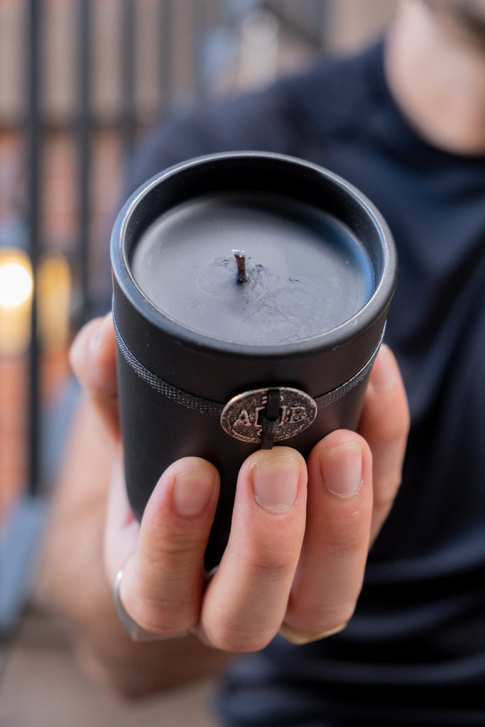 Someone holds the Archipelago - Black Forest 5.2oz Boxed Candle in a black jar with a single wick; its metal emblem and textured band hint at the rich Black Forest fragrance. The blurred background reveals dim lights and a railing, while the person wears a dark shirt.