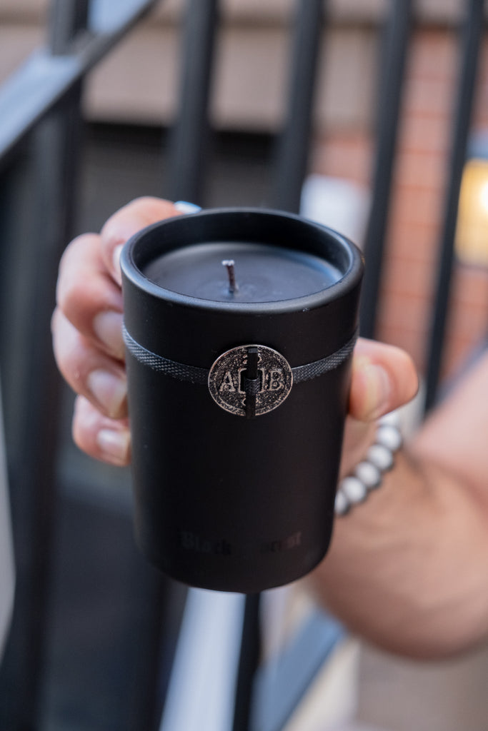 A person holds the Archipelago Black Forest 5.2oz Boxed Candle, featuring a matte black holder with a silver emblem and exuding the aroma of Dark Ebony Woods. They wear a beaded bracelet, while blurred railings and buildings in the background enhance the mysterious ambiance.