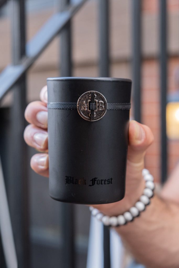 A hand holds the Archipelago Black Forest 5.2oz Boxed Candle with a textured silver band, emitting the Dark Ebony Woods fragrance. Brick walls and metal stairs blur in the background, while a white beaded bracelet adds elegance to the scene.