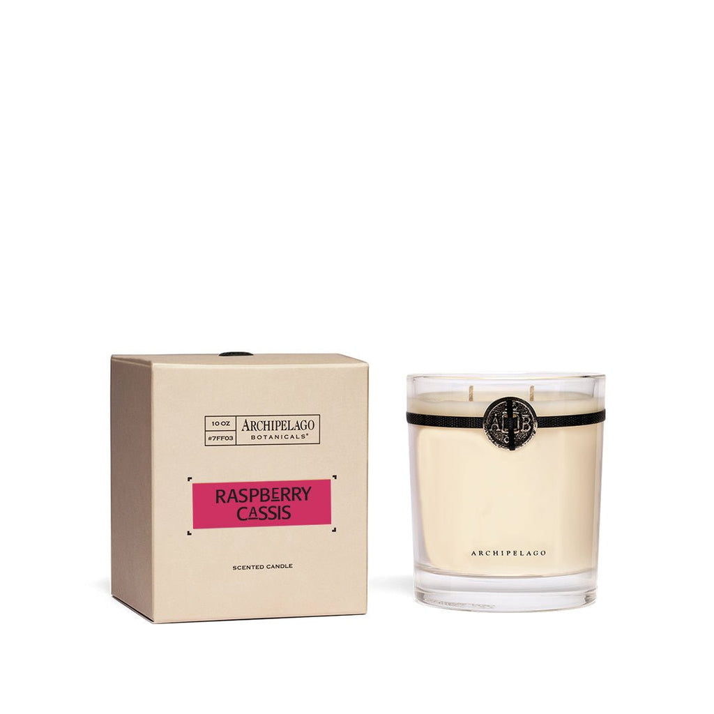 The Archipelago - Raspberry Cassis Credenza 10oz Candle, featuring a pink label and housed in clear glass with a black seal, sits next to its beige box. This summer-scented candle looks elegant against a white background, ideal for brightening any space.