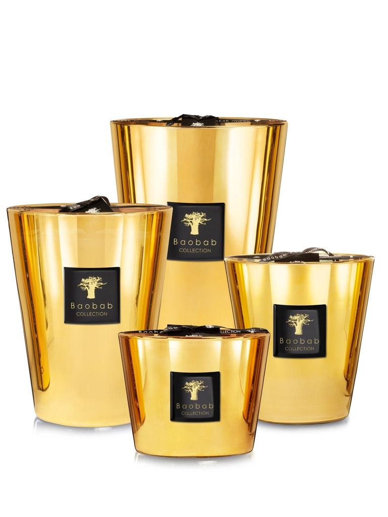 Four shiny, gold-colored Baobab Les Exclusives Aurum Max 10 candles are displayed. Each emits a delicate floral scent and features a distinctive black label with a tree logo. They vary in height, creating a visually appealing arrangement.