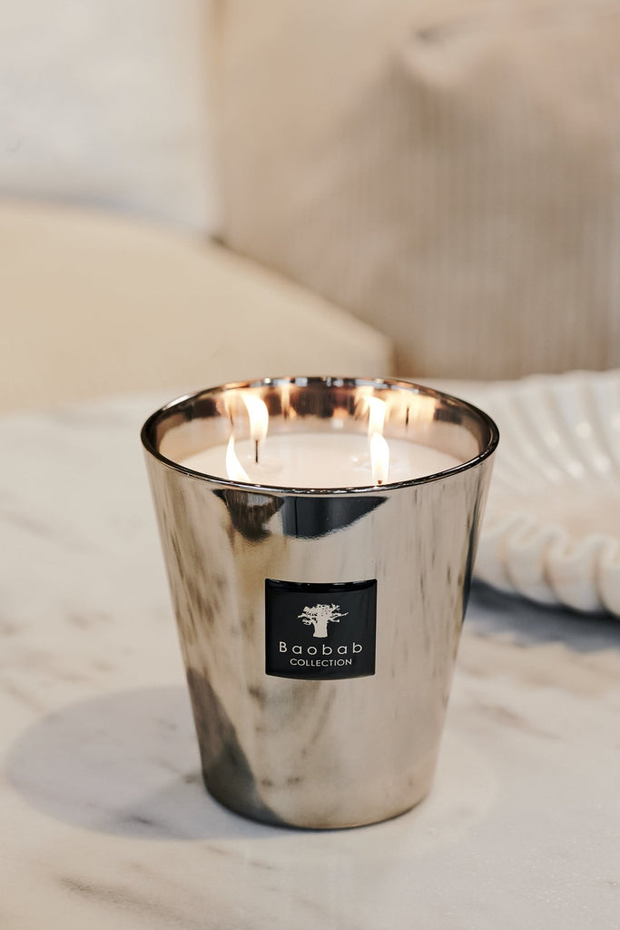 A Baobab candle from the Les Exclusives Platinum Max 16 collection, with a platinum scent and label, burns with two flames on a marble surface. The blurred, light-colored backdrop hints at a cozy room filled with an amber fragrance.