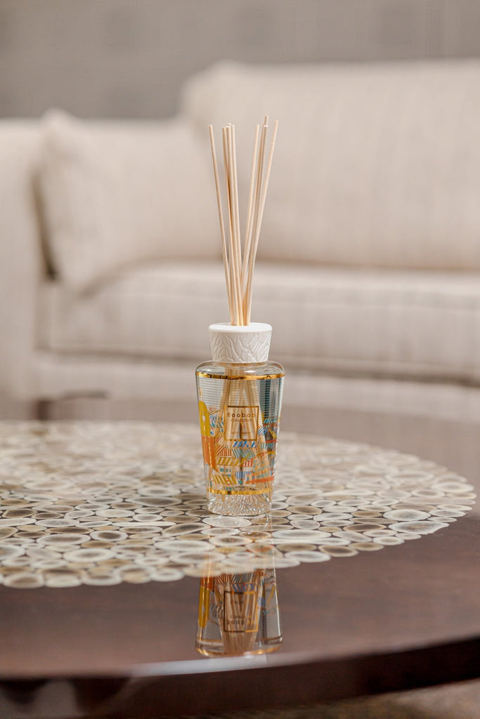 A Baobab - My First Baobab A Saint-Tropez 8.45oz Reed Diffuser sits on a polished wooden table, displaying an intricate circular design. Clear home fragrance with natural reeds adds elegance, while a cream-colored sofa forms the perfect backdrop to this harmonious scene.