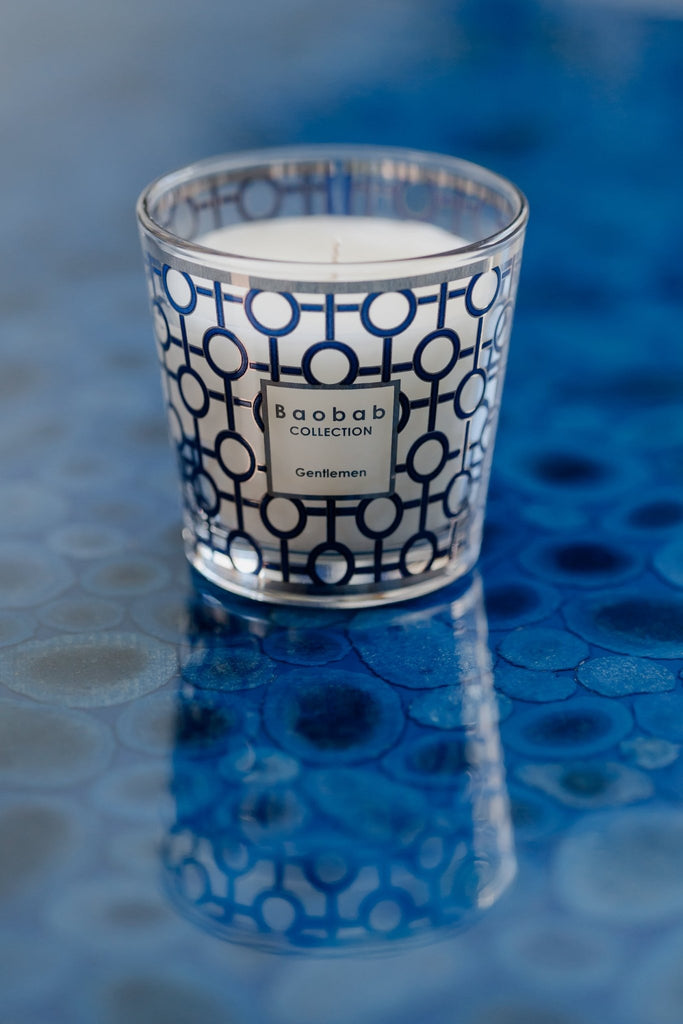 A lit Boabab - My First Baobab Gentlemen 6.7oz Candle in a geometric glass holder rests on a textured blue surface. Labeled Baobab, it suggests a masculine aroma, casting a gentle reflection on the table.