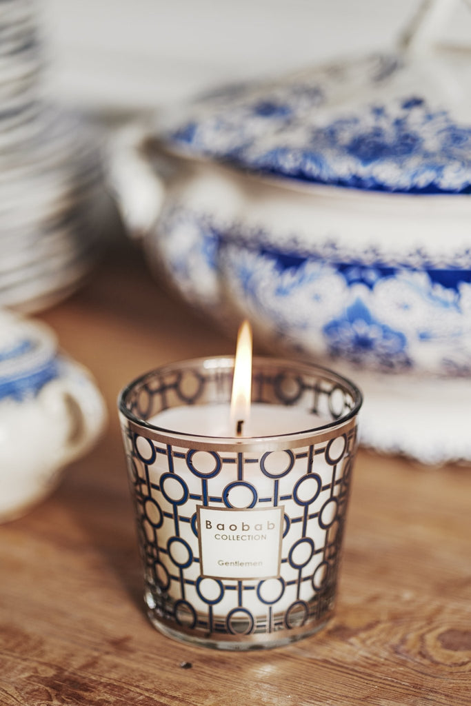 A 6.7oz Baobab candle, My First Baobab Gentlemen, in a decorative glass holder featuring a geometric pattern, rests on wood. Behind it lies blue and white patterned crockery with a lidded dish, embodying a refined masculine scent by the Baobab brand.