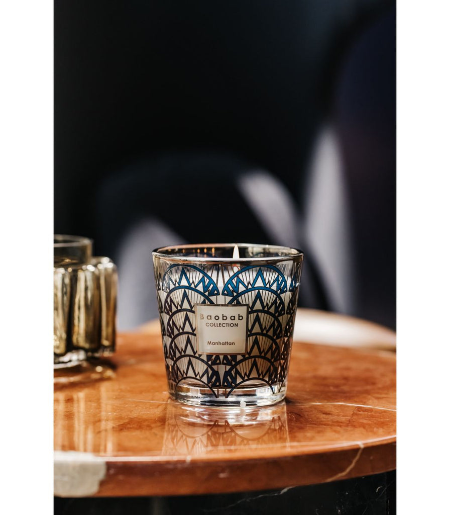 A Boabab - My First Baobab Manhattan 6.7oz Candle sits in a decorative glass holder on a wooden table. Featuring a geometric blue art deco design with the Baobab label, this piece adds elegance, while the blurred background enhances the cozy ambiance.