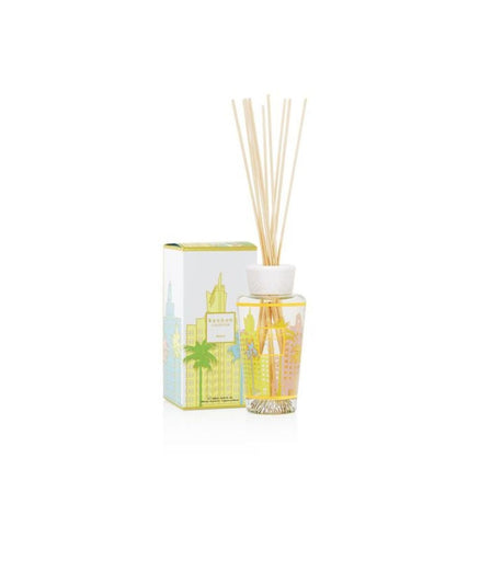 The Baobab - My First Baobab Miami 8.45oz Reed Diffuser has a clear glass bottle with yellow liquid and reed sticks. With an Art Deco fragrance, its packaging displays a colorful cityscape and palm trees, exuding the enchanting scent of Cypress Cedar.